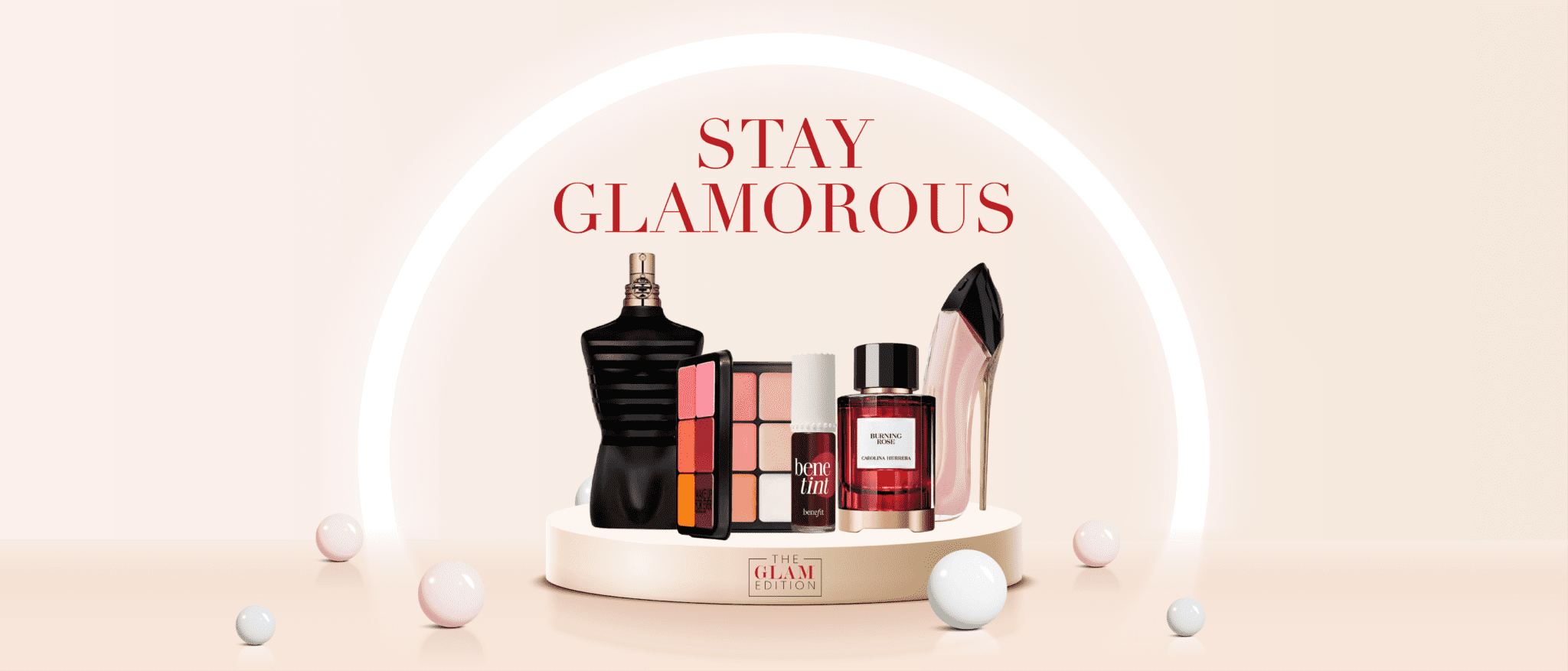 A beauty banner featuring luxury makeup and perfume products displayed on a round podium, with soft pink lighting, floating spheres, and the text ‘Stay Glamorous’ above them. Products include Jean Paul Gaultier perfume, a makeup palette, Benefit Benetint, Carolina Herrera Burning Rose, and a stiletto-shaped perfume bottle, with The Glam Edition logo at the base.
